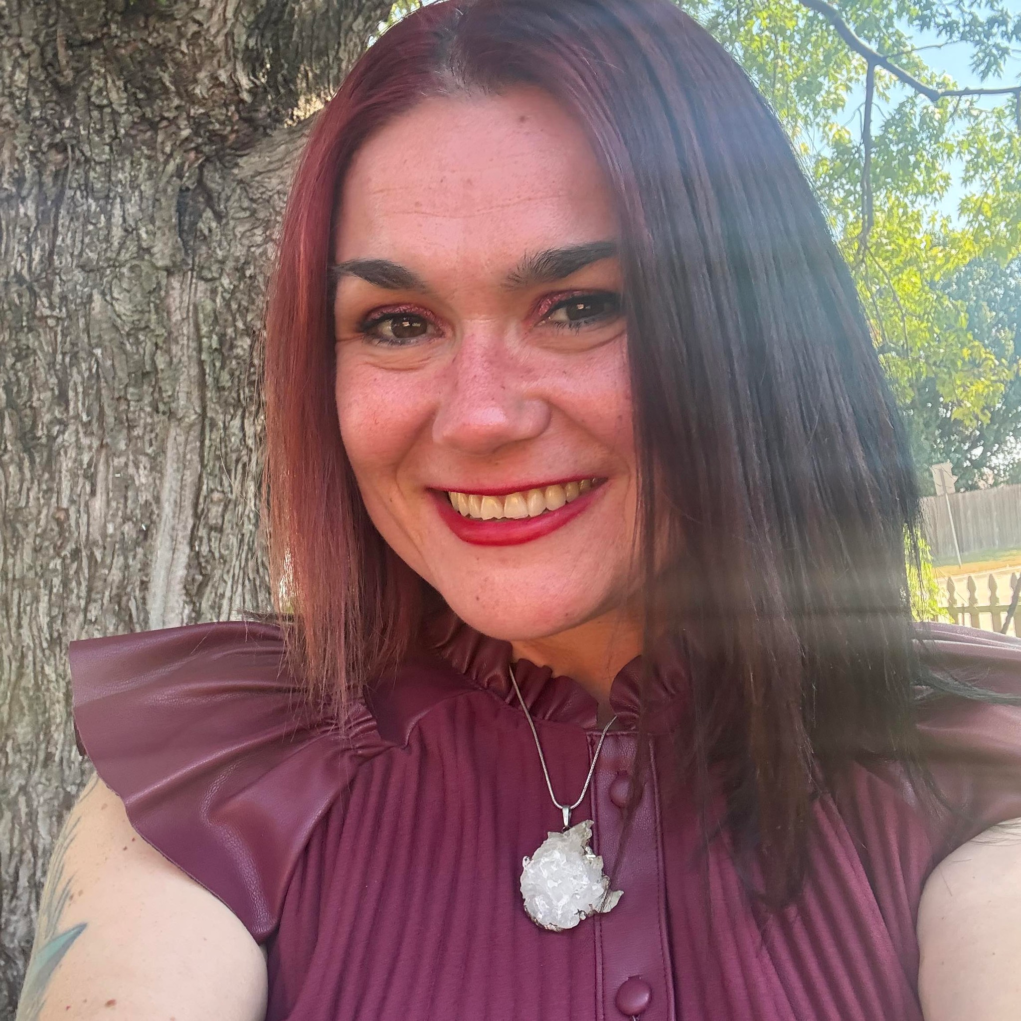 A smiling woman with straight dark red hair and makeup, wearing a maroon top with ruffled shoulders and a necklace with a large white crystal pendant, standing outdoors near a tree with a fence and green trees in the background.