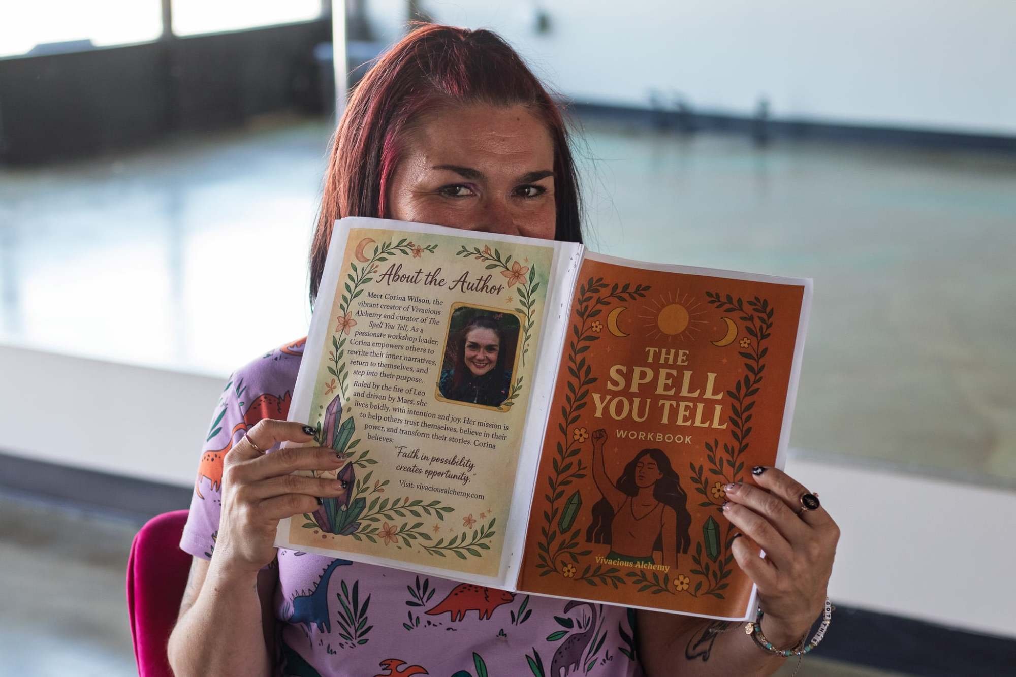 Woman holding up a book titled 'The Spell You Tell' Workbook, partially covering her face, inside a room with large windows.