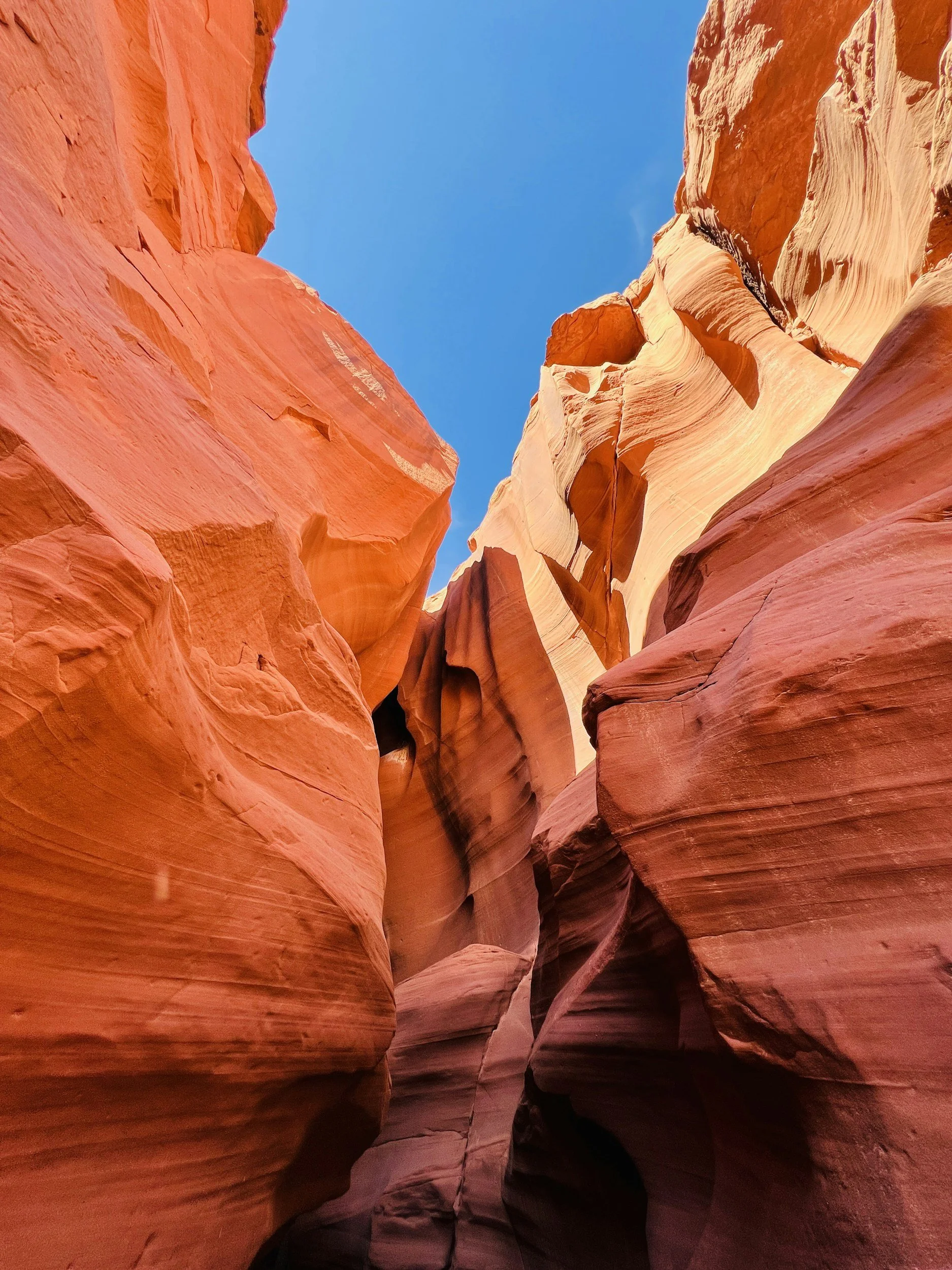 Tall, narrow slot canyon with smooth, layered orange and pink sandstone walls reaching towards a bright blue sky.