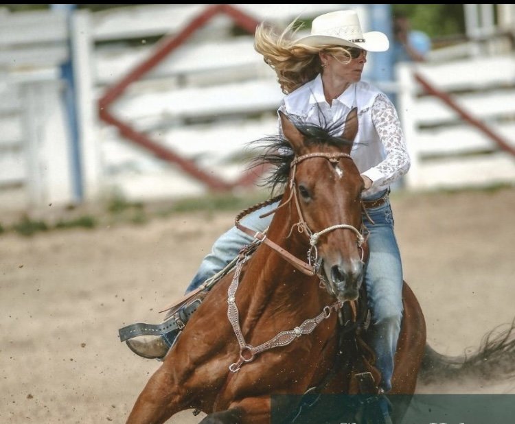 A woman wearing a white cowboy hat and sunglasses, riding a galloping brown horse on a dirt track in an outdoor rodeo arena.