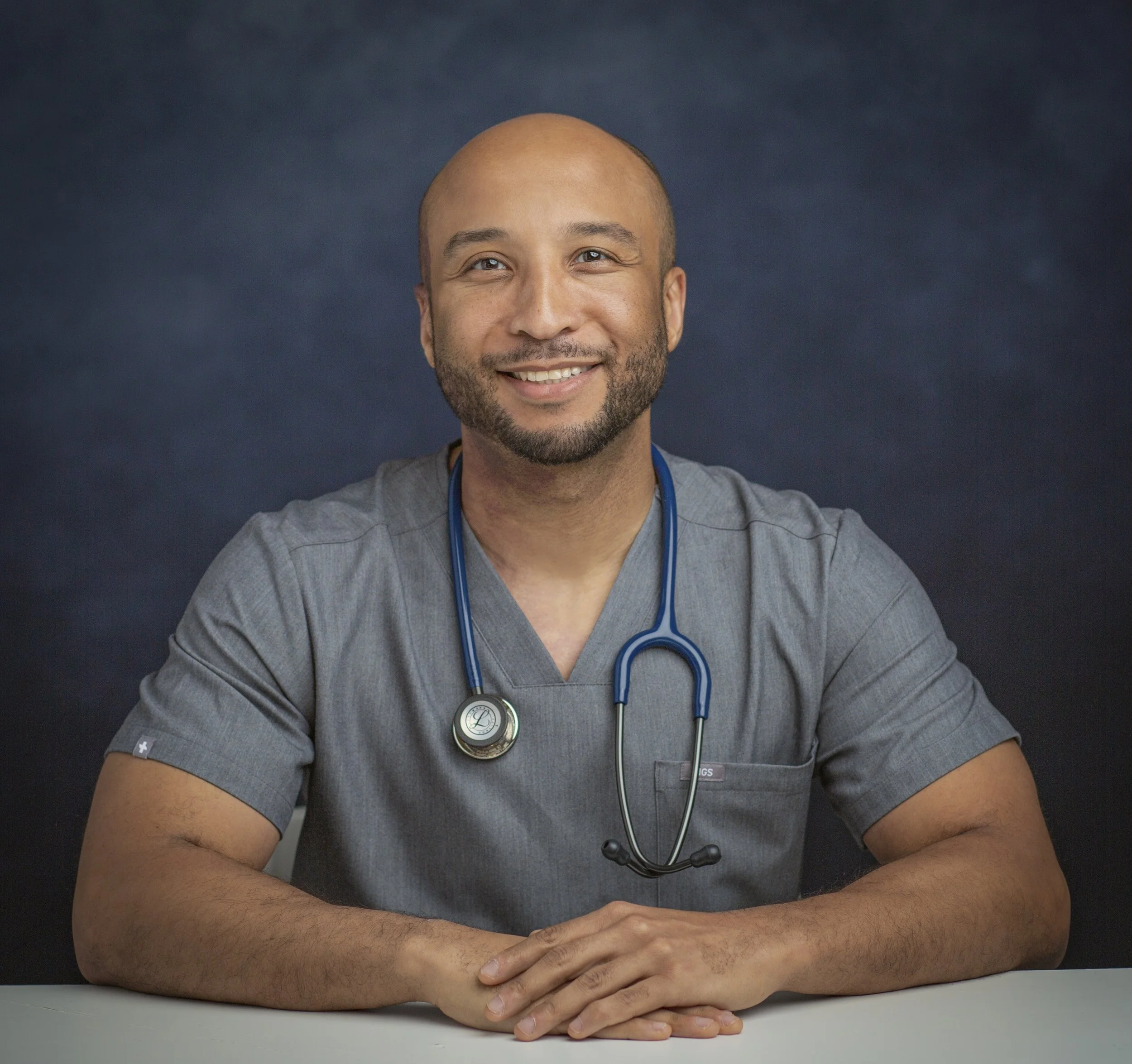 A male healthcare professional with a stethoscope smiling at the camera, seated at a table against a dark background.