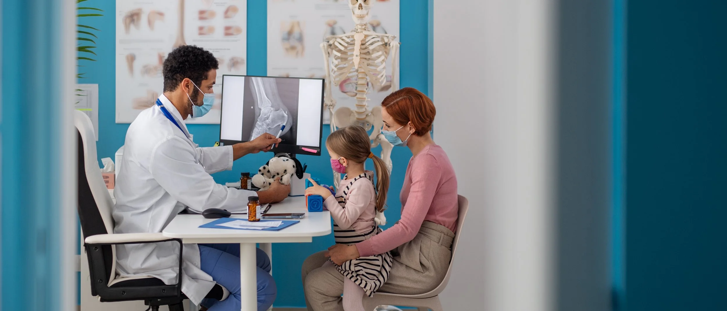 A pediatric doctor explaining an X-ray of an ankle to a young girl and her mother in a medical examination room. The doctor is seated, pointing at the X-ray on a monitor, with medical posters and a skeleton model in the background. The girl and mother are wearing masks, engaging with the doctor.