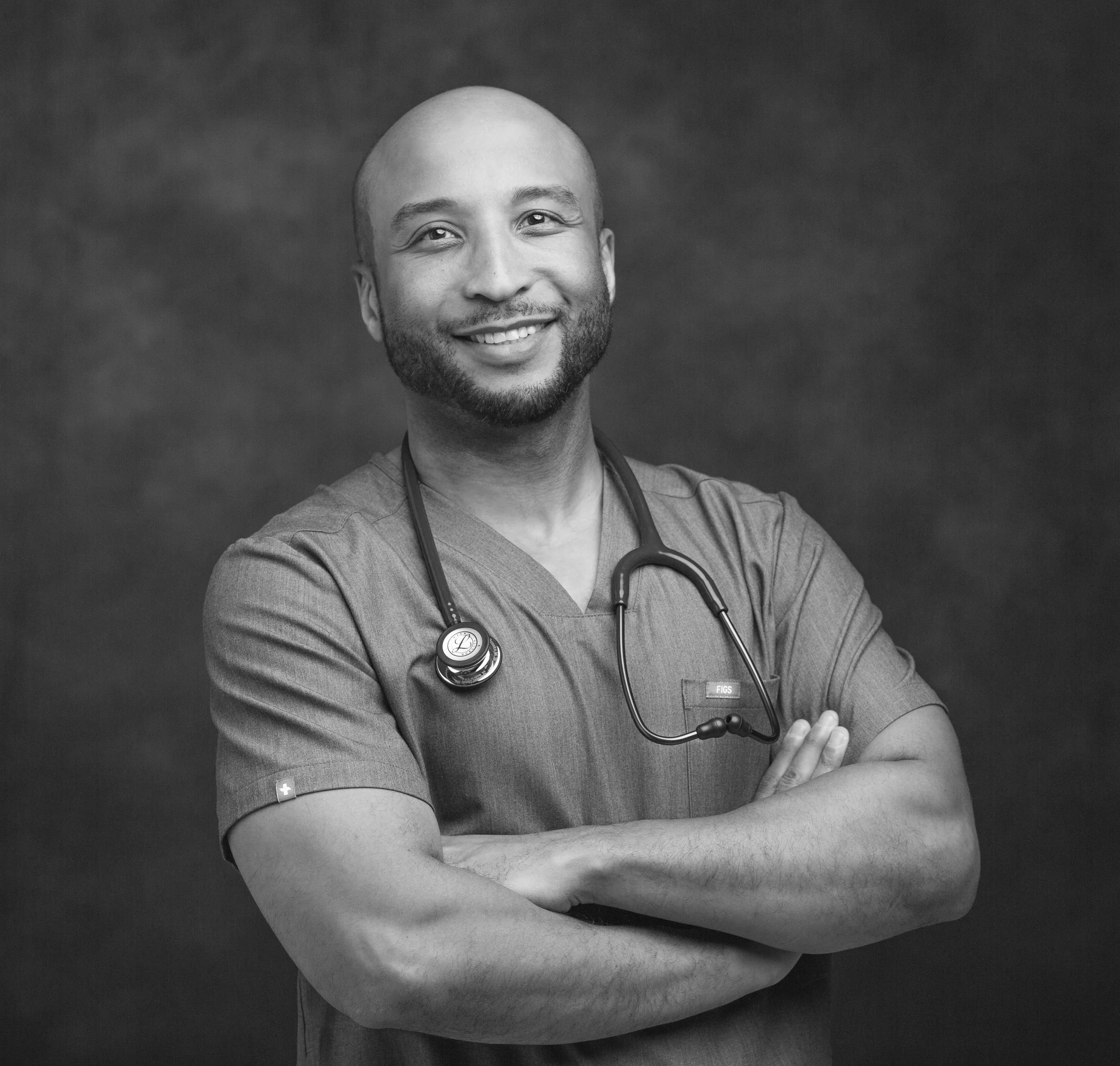 A smiling male healthcare professional with a beard and stethoscope around his neck, wearing medical scrubs, standing against a dark background.
