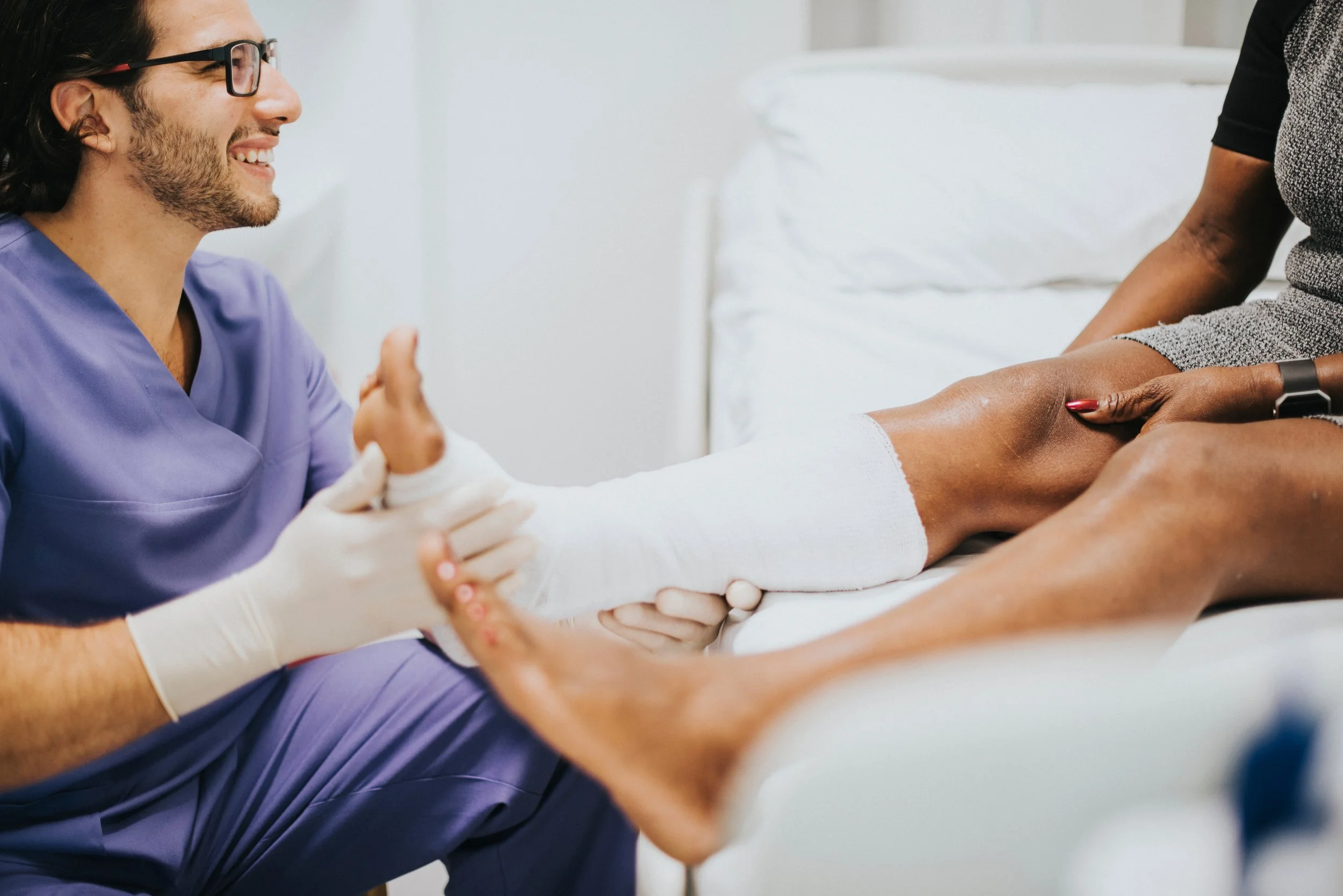 A medical professional in scrubs is holding and examining a patient's leg, who is sitting on a hospital bed in a medical setting.