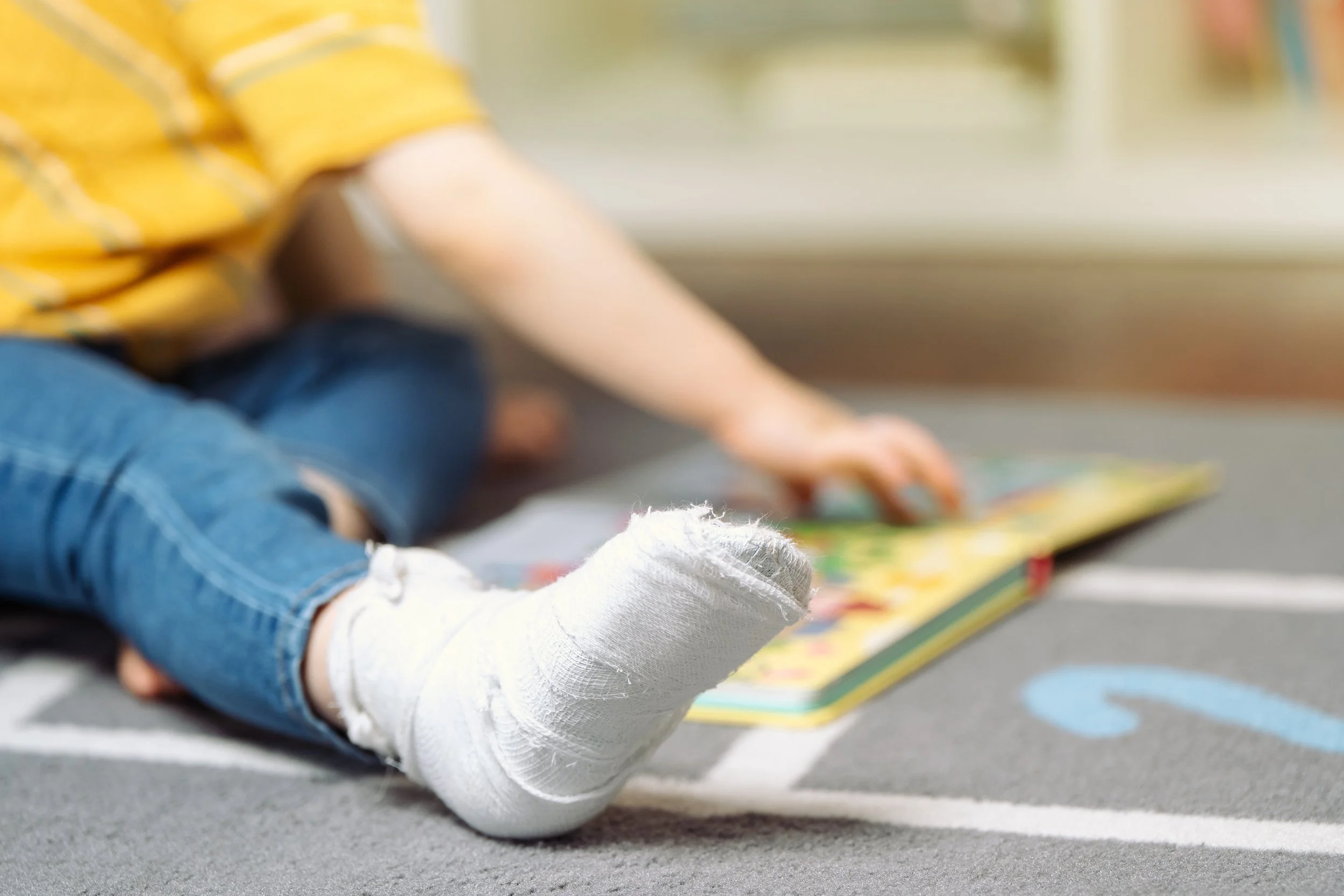 Child with a broken ankle in a cast, sitting on the floor reading a book.