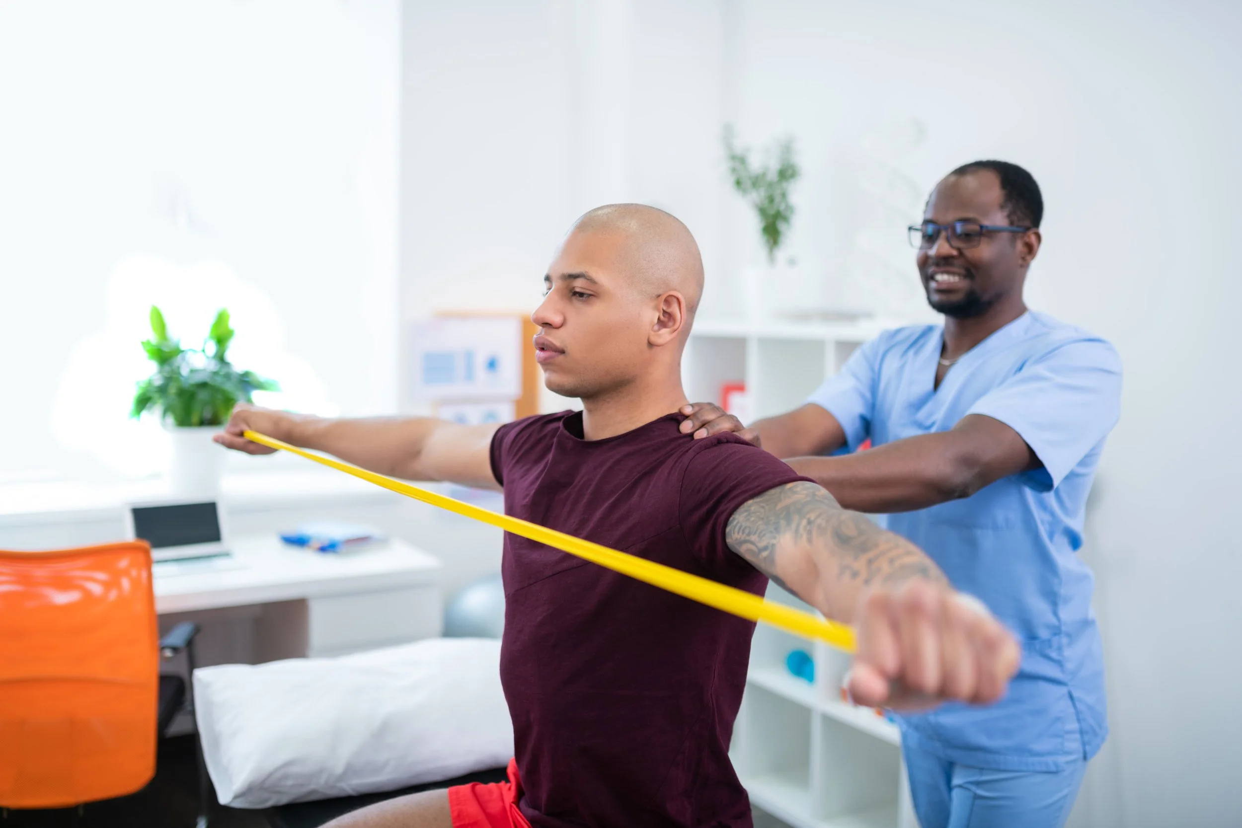 A physical therapist assists a young man with shoulder stretches using a yellow resistance band in a bright therapy room.