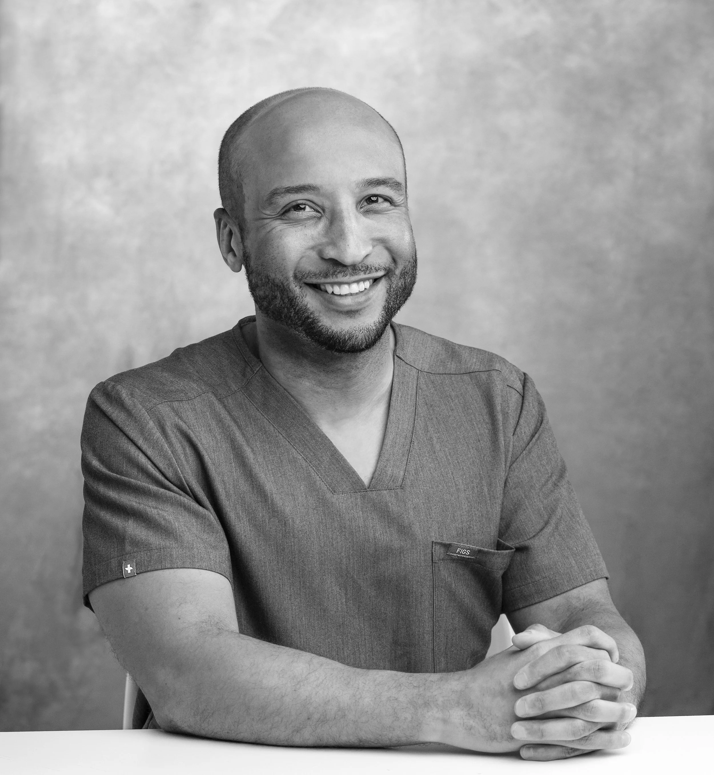 Black-and-white portrait of a smiling male healthcare professional in scrubs sitting at a table with hands clasped.