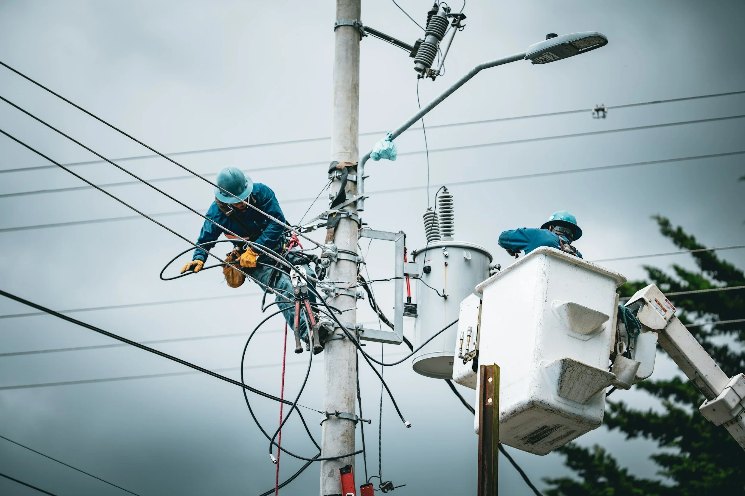 Two utility workers in safety gear repairing electrical wires on a utility pole during overcast weather.