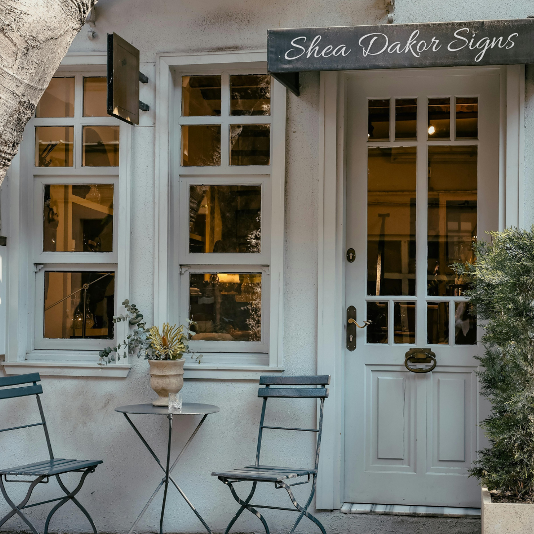 storefront with a sign reading 'Shea Dakar Signs'; outdoor seating with two black chairs and a small table with a potted plant.