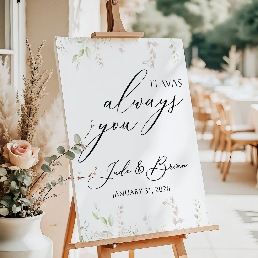 Wedding reception sign with floral design reading 'It was always you Job & Brian January 31, 2026' on an easel, with flower arrangements nearby and a softly lit indoor setting.