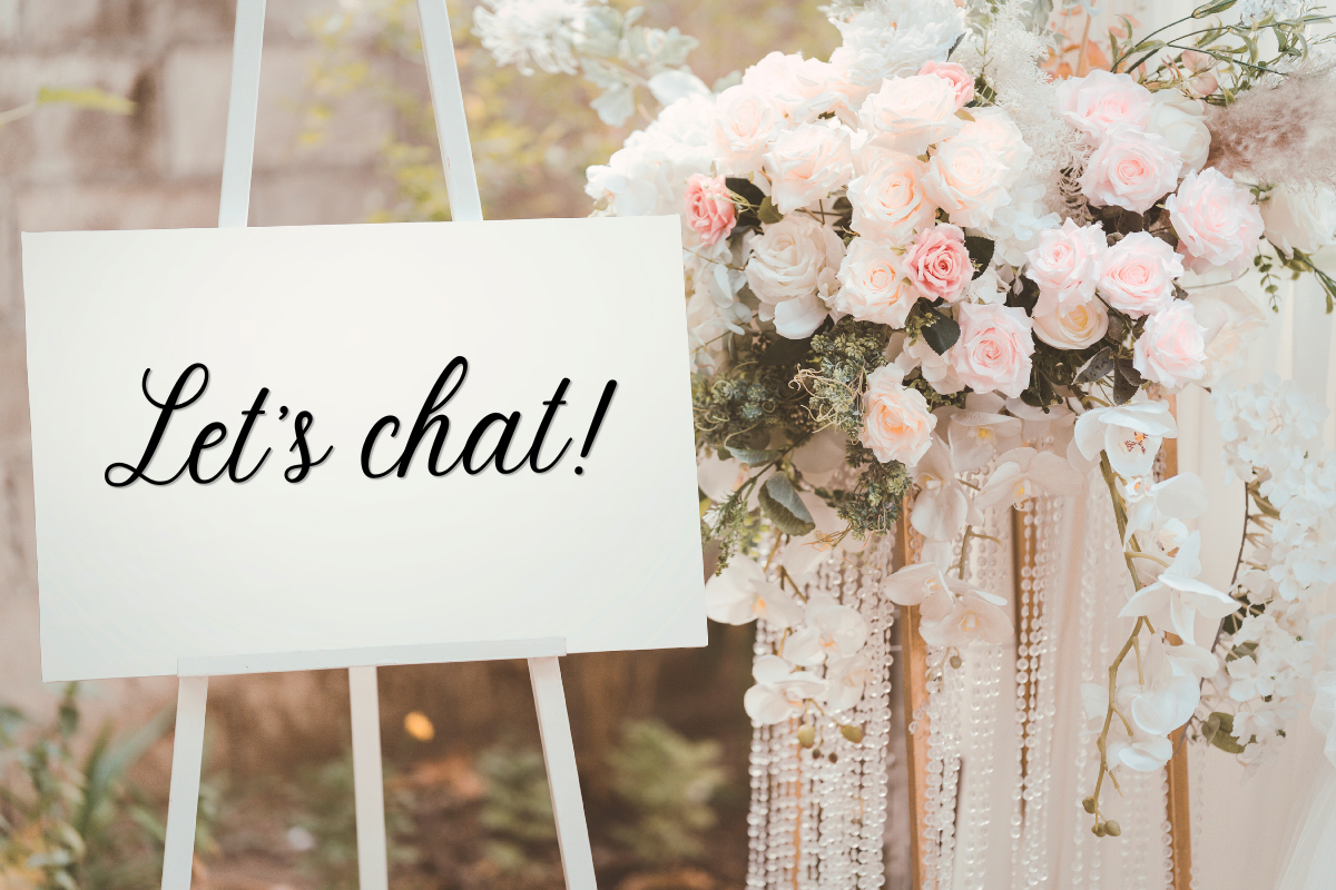 A white signboard with the text 'Let's chat!' written in black cursive font, placed on an easel beside a floral arrangement of pink and white roses and white orchids, with a blurred outdoor garden background.