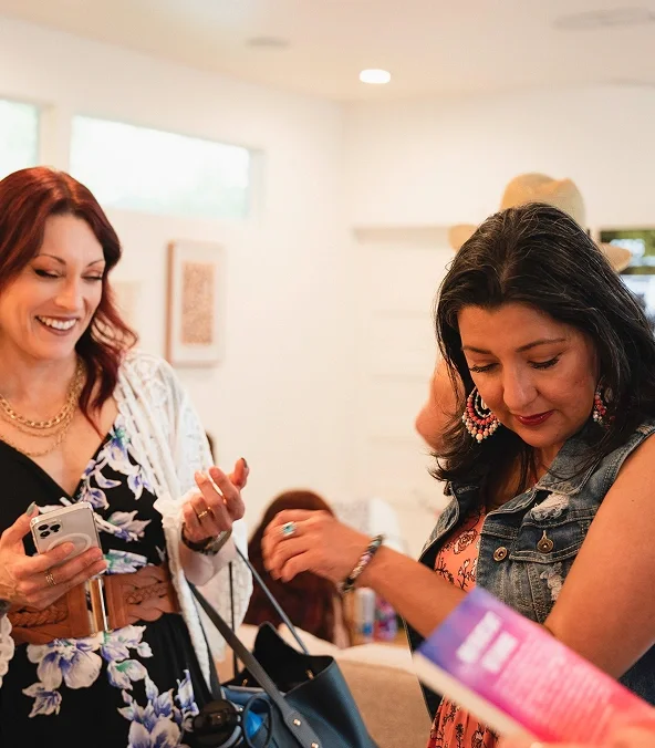 Two women smiling and looking at their phones at a social gathering indoors.