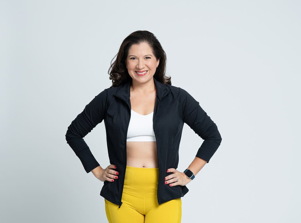 Bold Mama, Melissa Acosta, with dark hair smiling, wearing a black jacket, white workout top, yellow leggings, and a smartwatch, standing with hands on hips against a plain gray background.