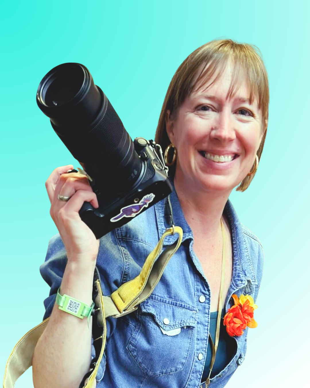 A smiling woman with short brown hair wearing a denim jacket, gold hoop earrings, and a yellow flower pin, holding a professional camera with a large telephoto lens against a colorful blue-green gradient background.