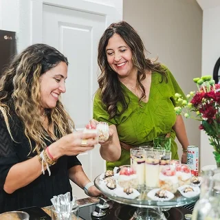 Two women sharing a dessert platter with cakes and drinks, smiling and enjoying their time together in a kitchen.