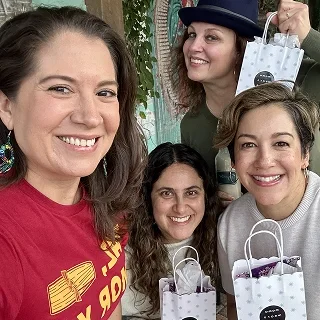 Four women smiling and holding shopping bags, standing outdoors near a tree.