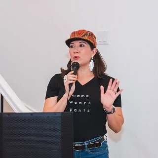 Woman, Melissa Acosta, speaking into a microphone, wearing a colorful cap, black t-shirt, and large earrings, standing in front of a white wall.
