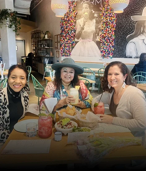 Three women sitting at a table in a restaurant, smiling with drinks and food. The background has a large black and white photo of a woman singing, decorated with colorful wall art and hanging lights.