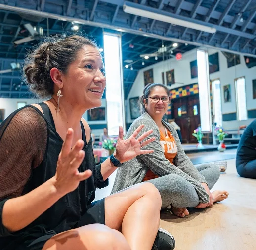 Two women sitting on the floor in a fitness place, engaged in conversation, with paintings and vibrant decor in the background.