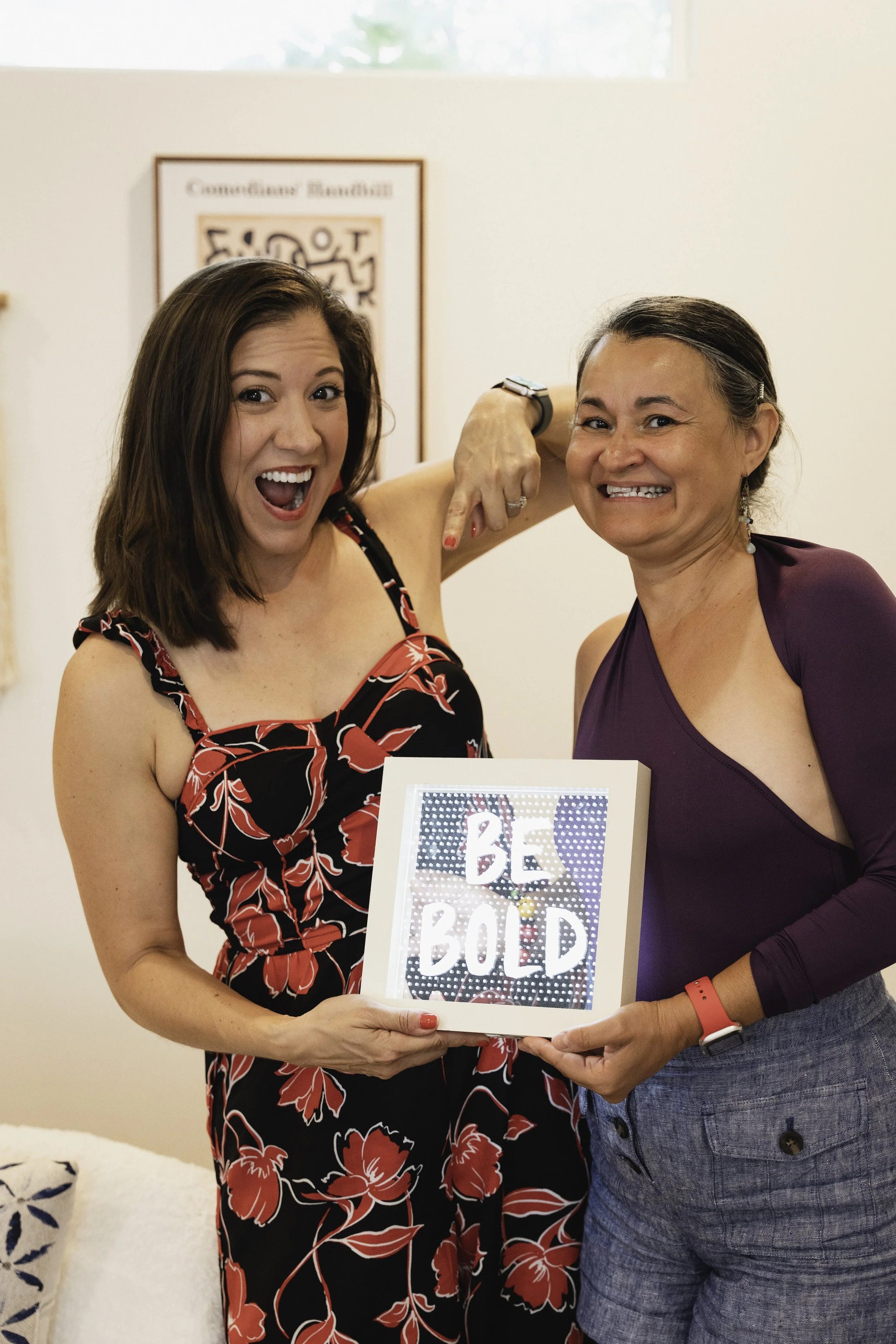 Two women smiling and holding a framed sign that says 'BE BOLD' in a room with artwork in the background.