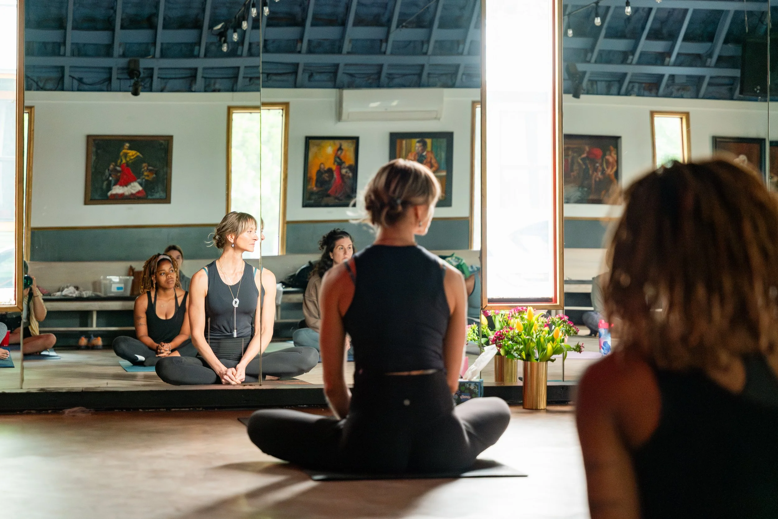Women practicing yoga in a studio with painted walls and a large mirror, with flowers placed near the mirror.