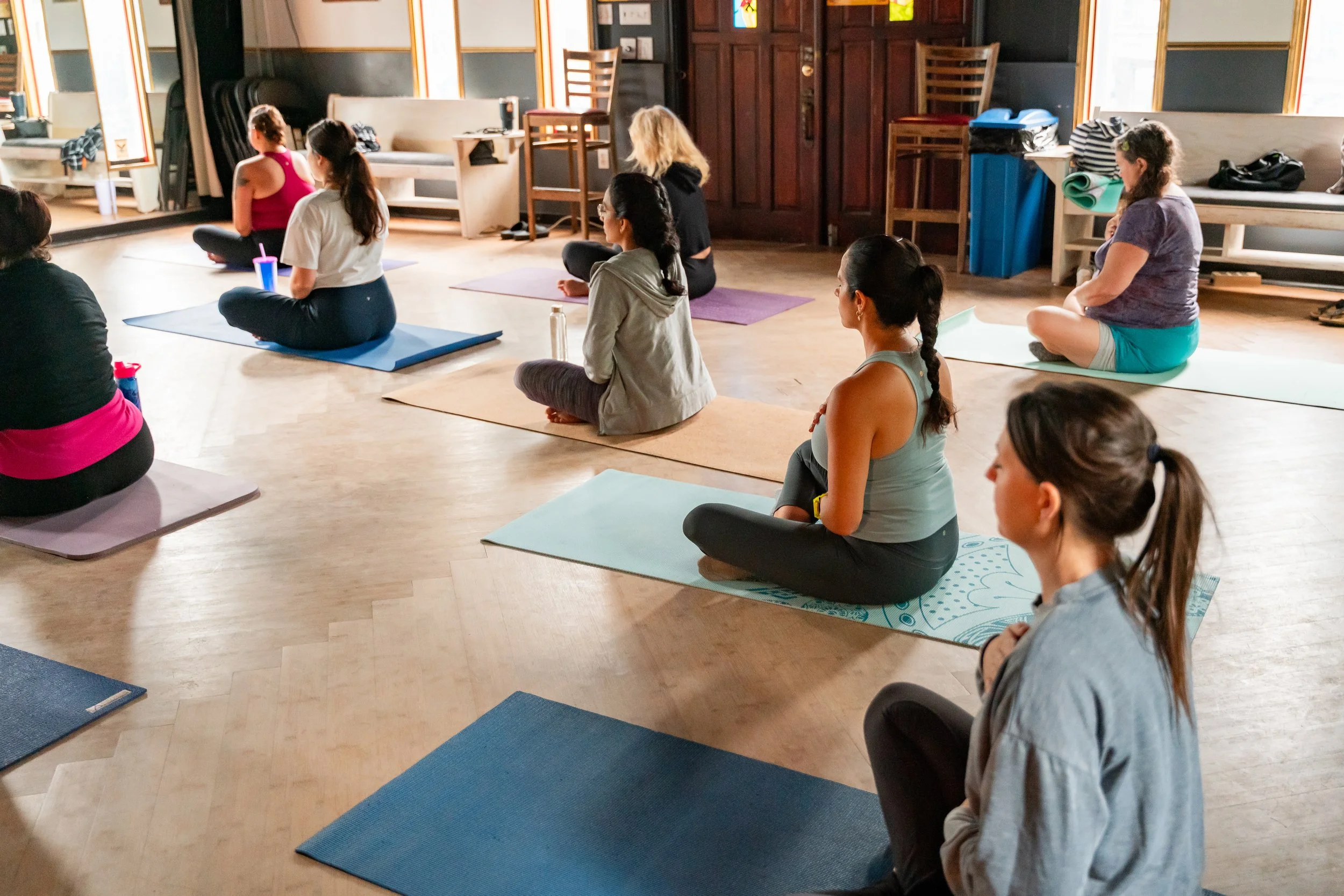 Group of women attending a yoga or meditation class in a studio, sitting cross-legged on mats, facing forward with closed eyes or relaxed expressions.