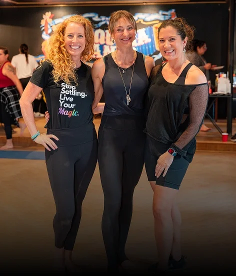 Three women standing together, smiling, in a dance or fitness studio with a dark background and neon lighting.