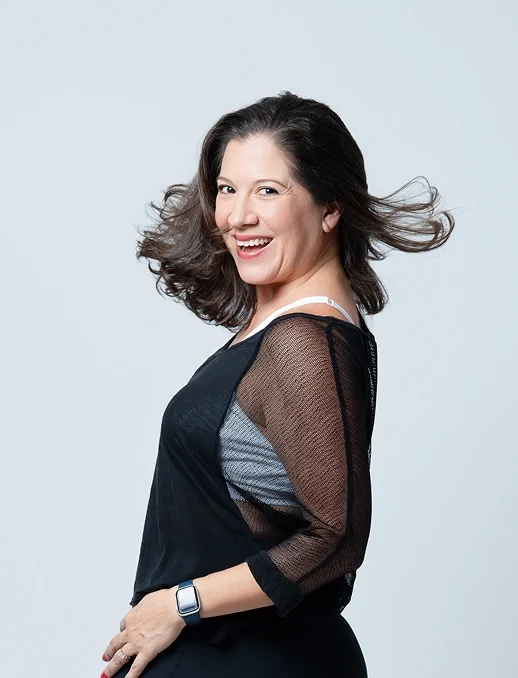 A  bold woman, Melissa Acosta, with shoulder-length brown hair, smiling and looking over her shoulder, wearing a black top with sheer sleeves and a smartwatch.