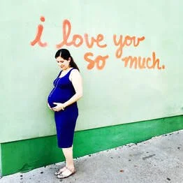 Melissa, pregnant, wearing a blue dress standing in front of a wall with the words 'I love you so much' written in orange.