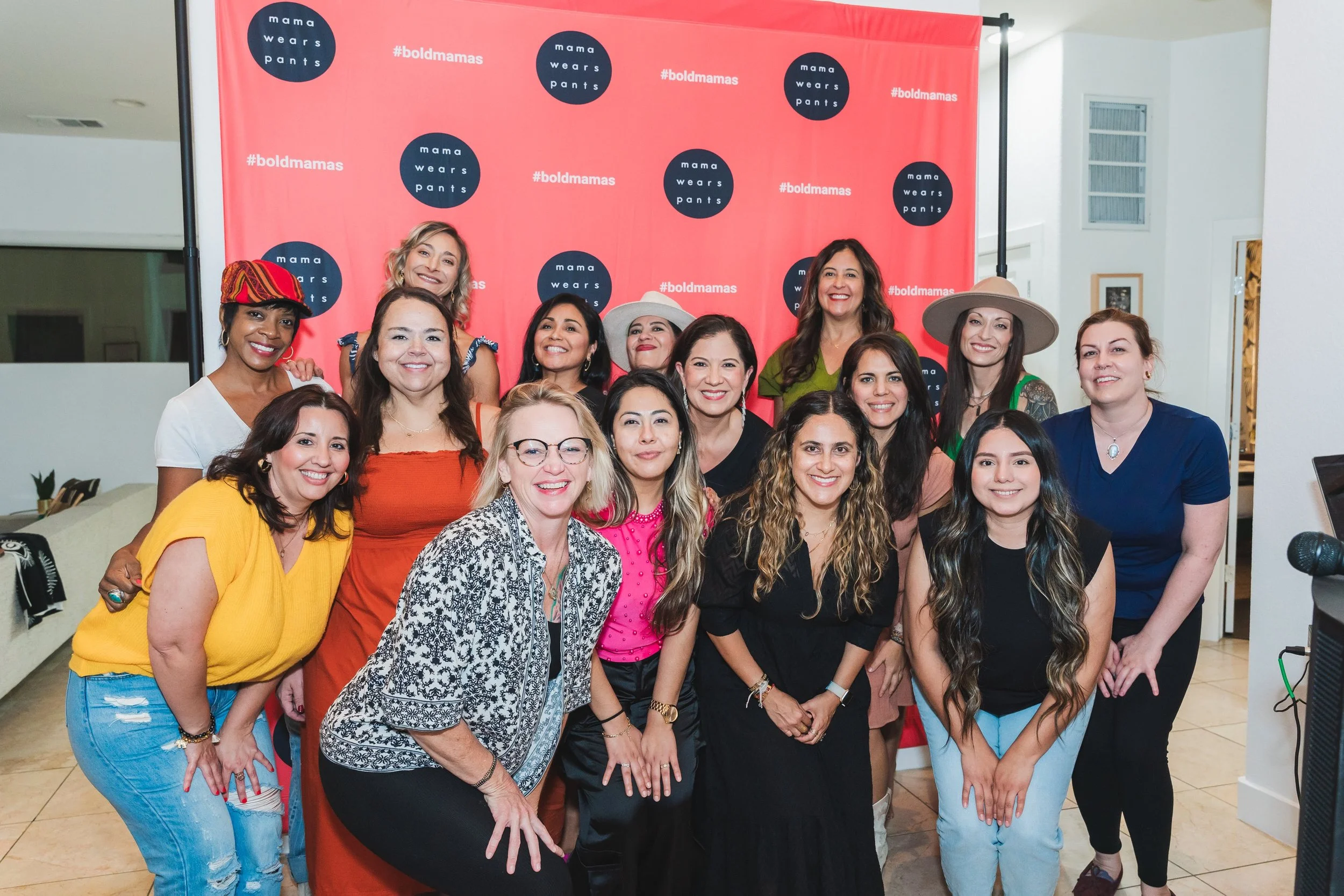 Group of women posing together indoors in front of a red backdrop with the text '#boldmamas' and 'mama wears pants' printed on it. The women are smiling and dressed casually, with some wearing hats.