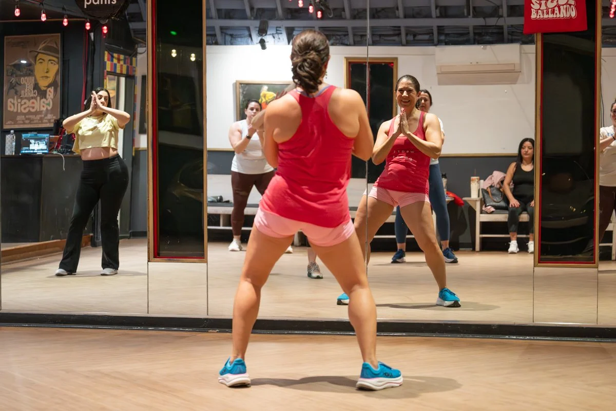 A group of women in a dance or fitness class, with the instructor facing a mirror and the participants watching her reflection. The instructor is in the foreground, wearing a red tank top and pink shorts, leading the class. The studio has a wooden floor, and the background shows wall art, a mirror, and some seated women.