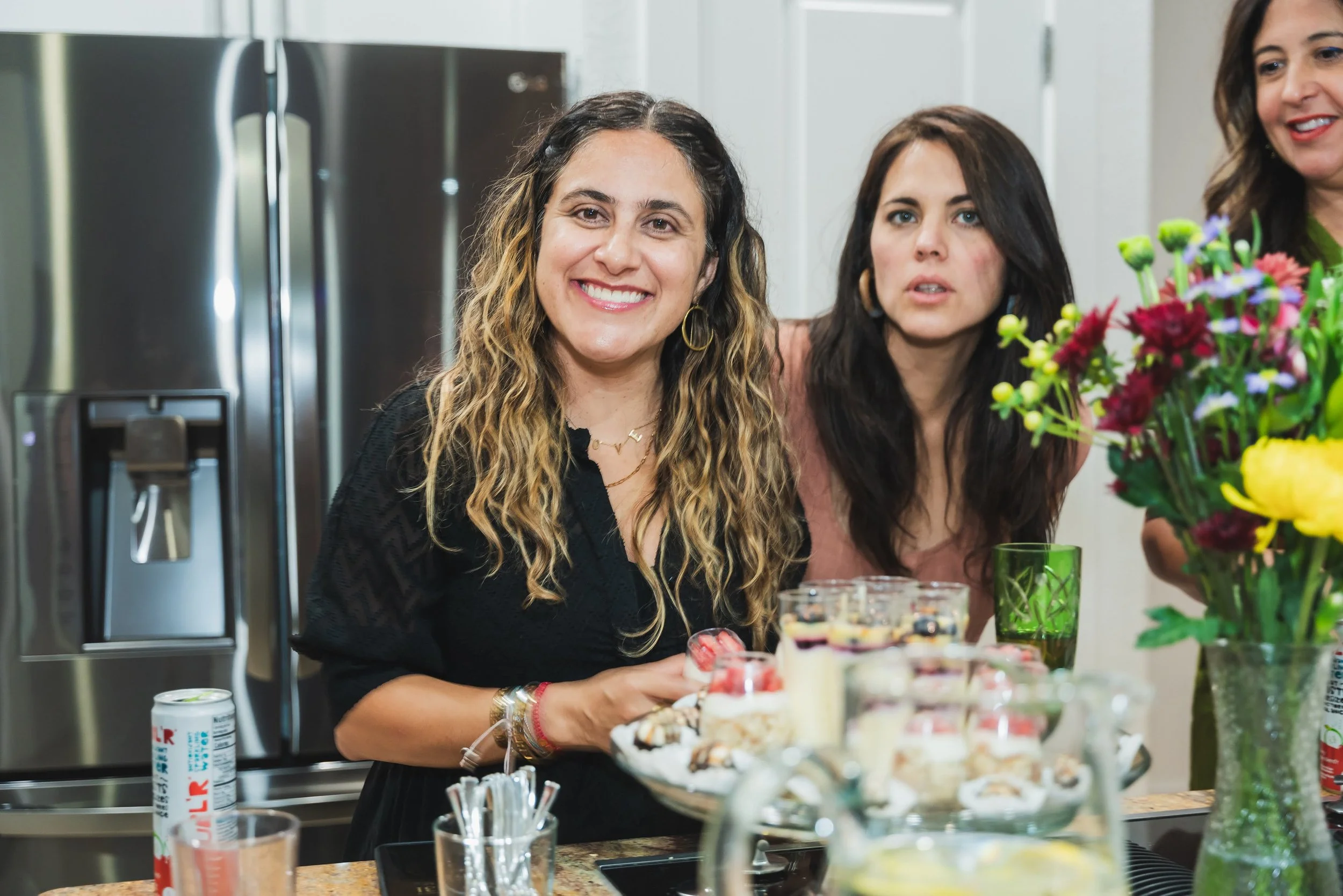 Three women at a gathering, one smiling, one looking curious, with colorful flowers and desserts on the table.