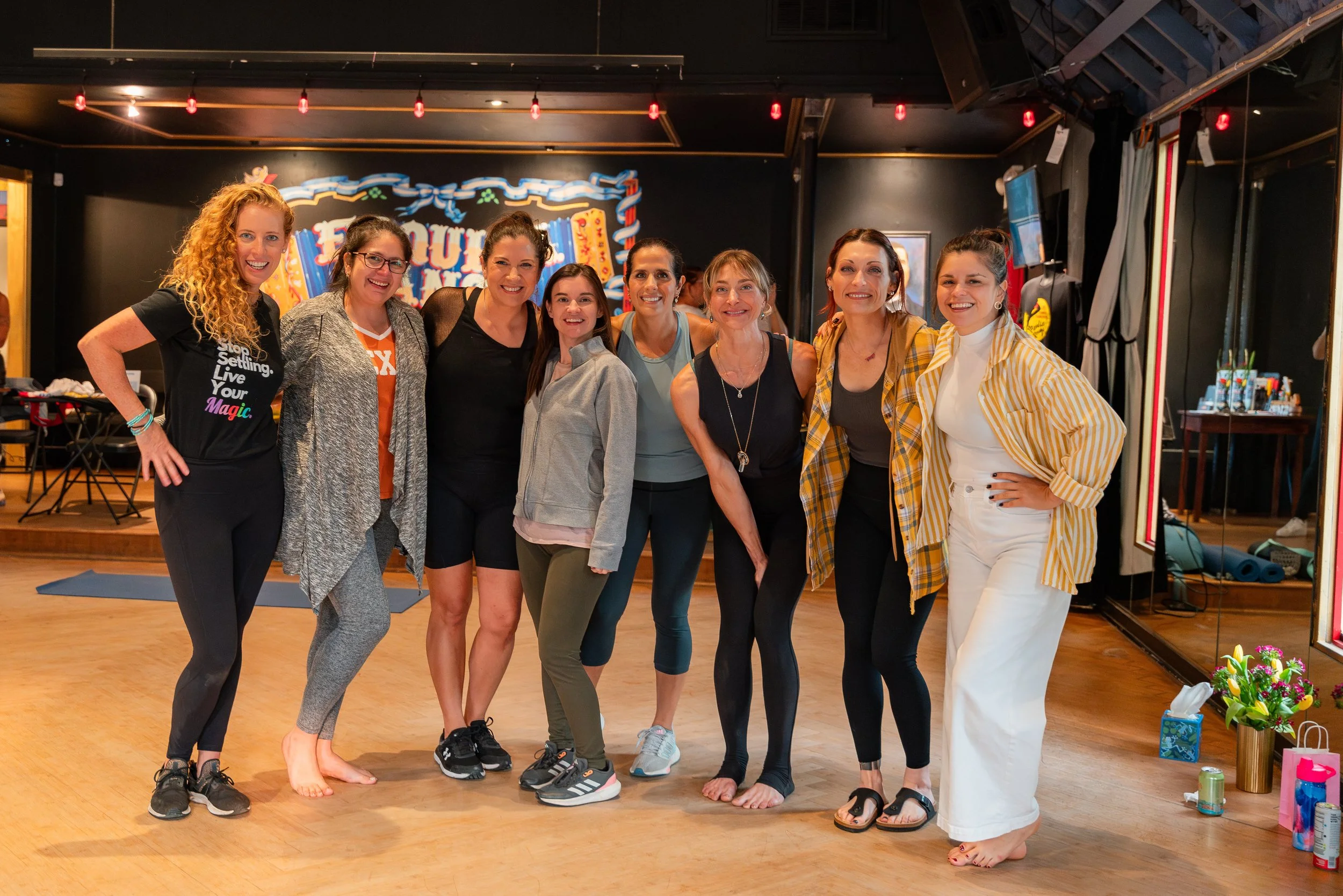 A group of eigth women posing together indoors on a wooden floor with a dark wall background, some wearing athletic attire, smiling and standing close together.