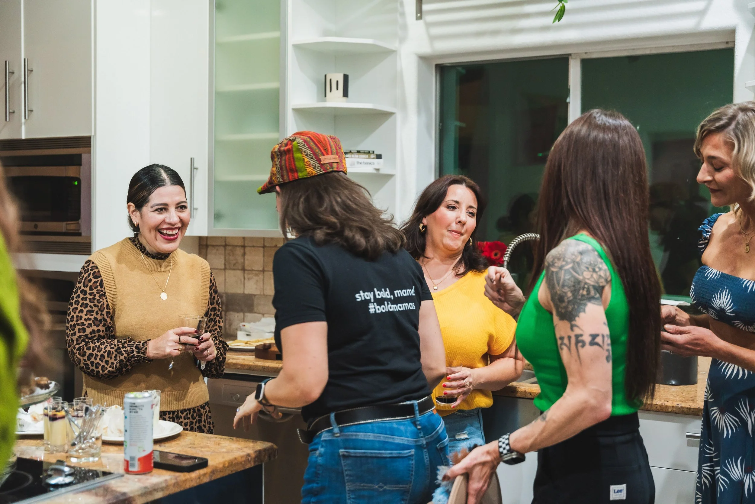 Group of women socializing in a kitchen, some holding drinks, engaging in conversation and smiling.