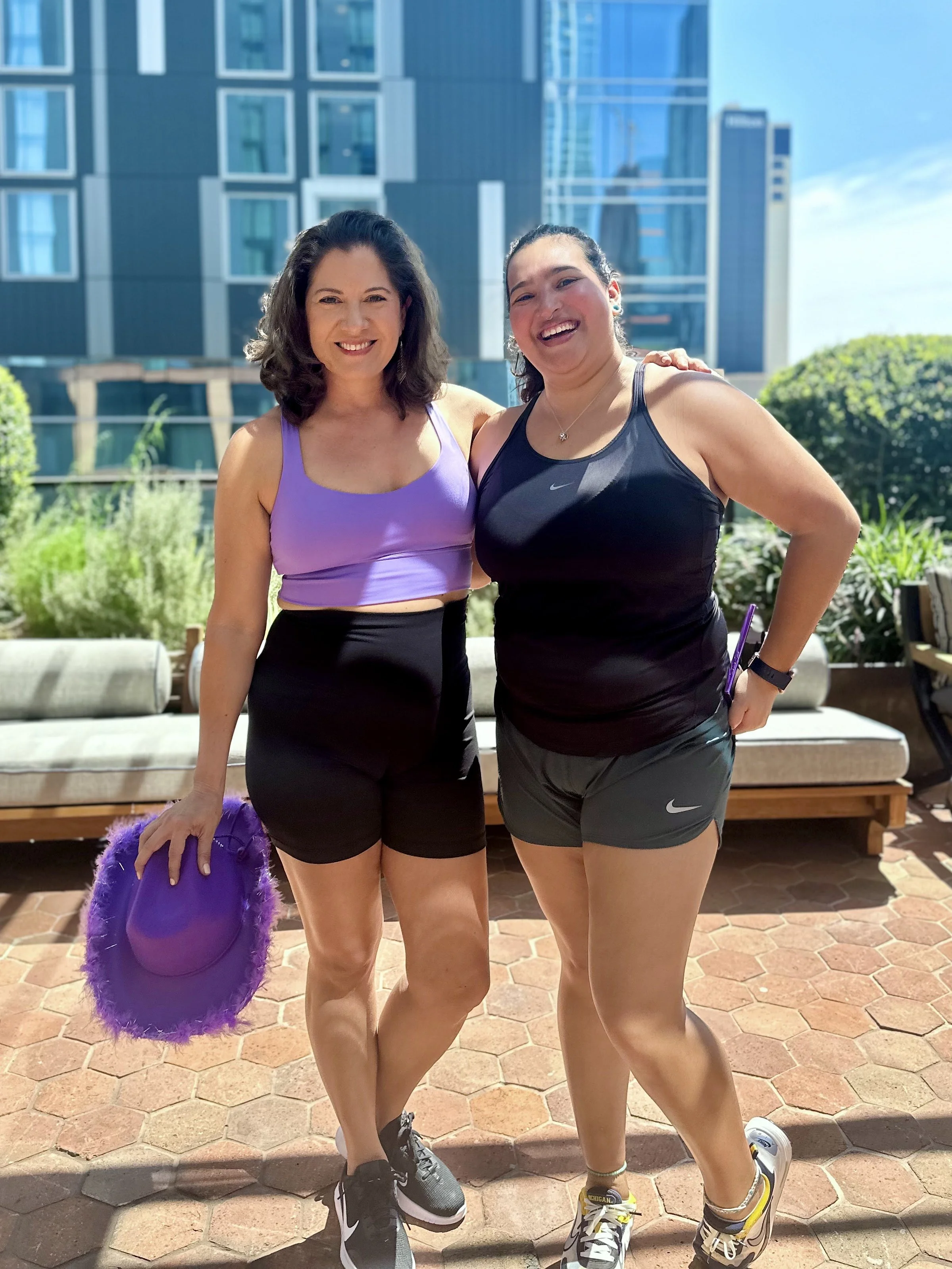 Two women in athletic clothing smiling outdoors with a modern building and greenery in the background.