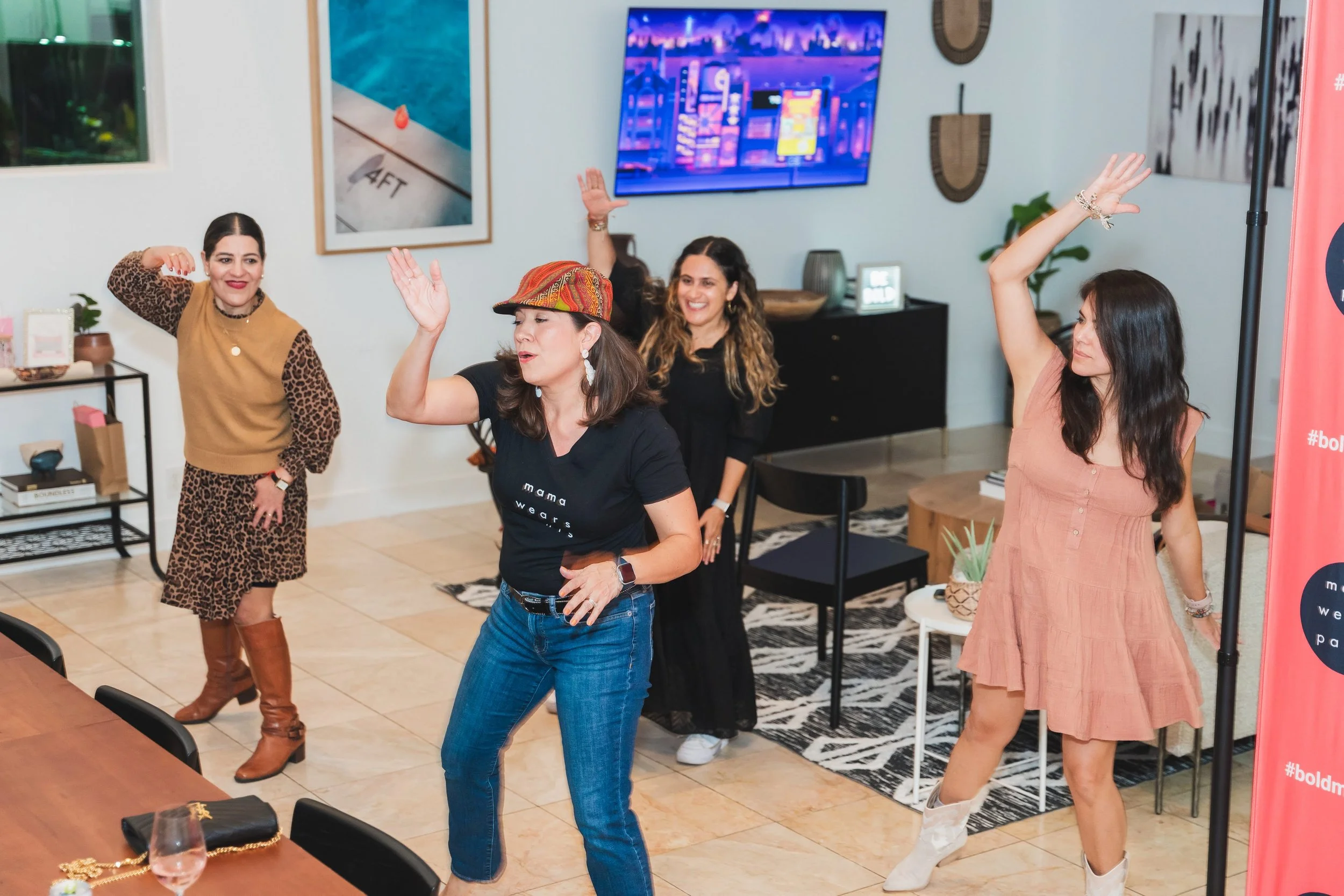 Four women dancing and having fun at a social gathering in a living room, with a large TV on the wall and decor accents.
