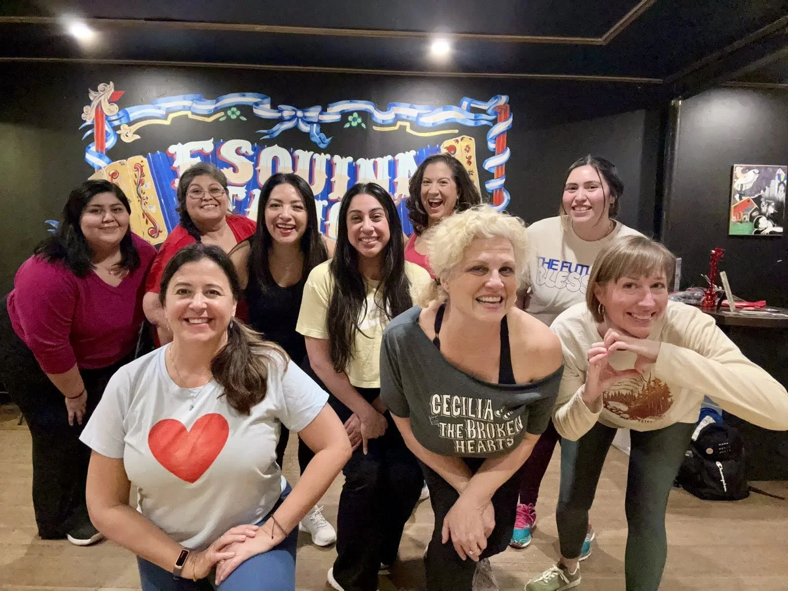A group of nine women smiling and posing for photo indoors, standing in front of a colorful mural with the text "Esquina Tango" in a lively setting that appears to be a fitness studio or zumba venue.