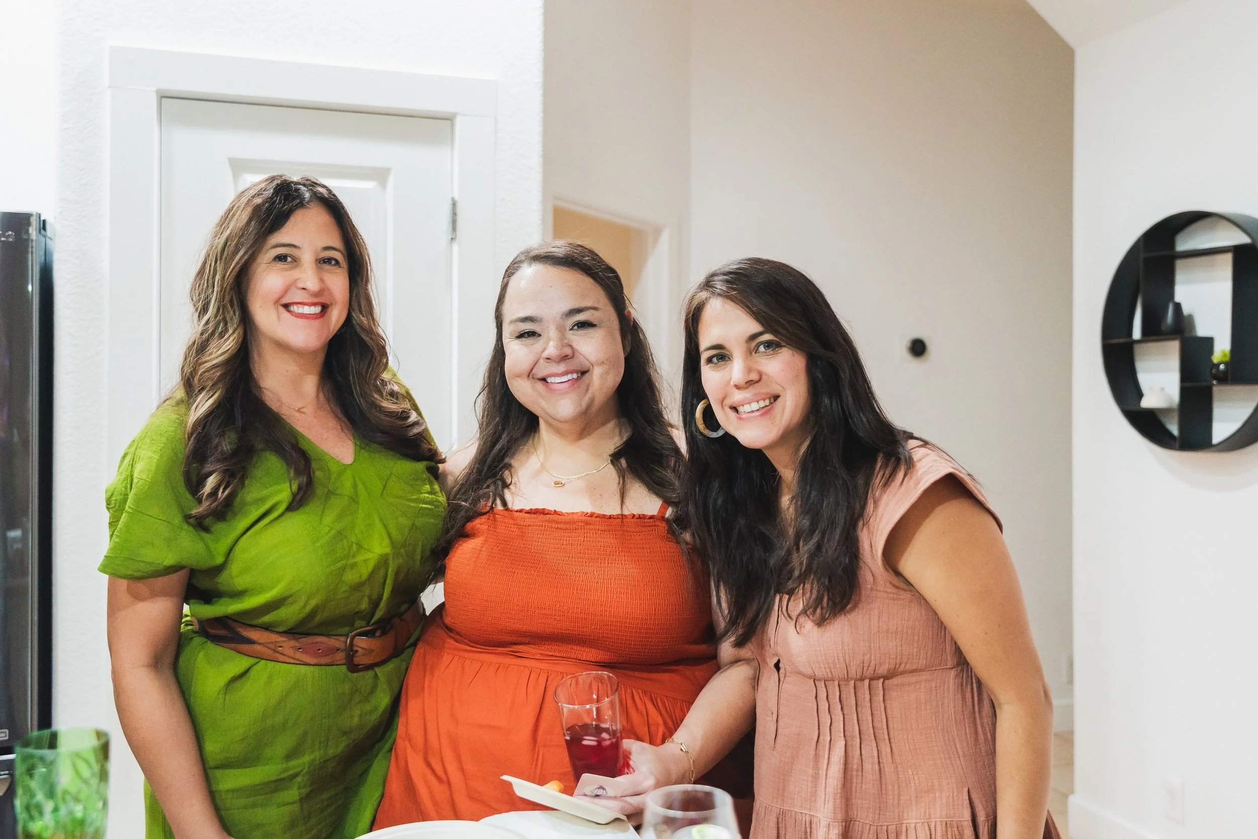 Three women smiling at a social gathering, standing together indoors. One woman on the left wears a green dress, the middle woman in an orange dress, and the woman on the right in a rust-colored dress. One woman holds a glass of red drink.