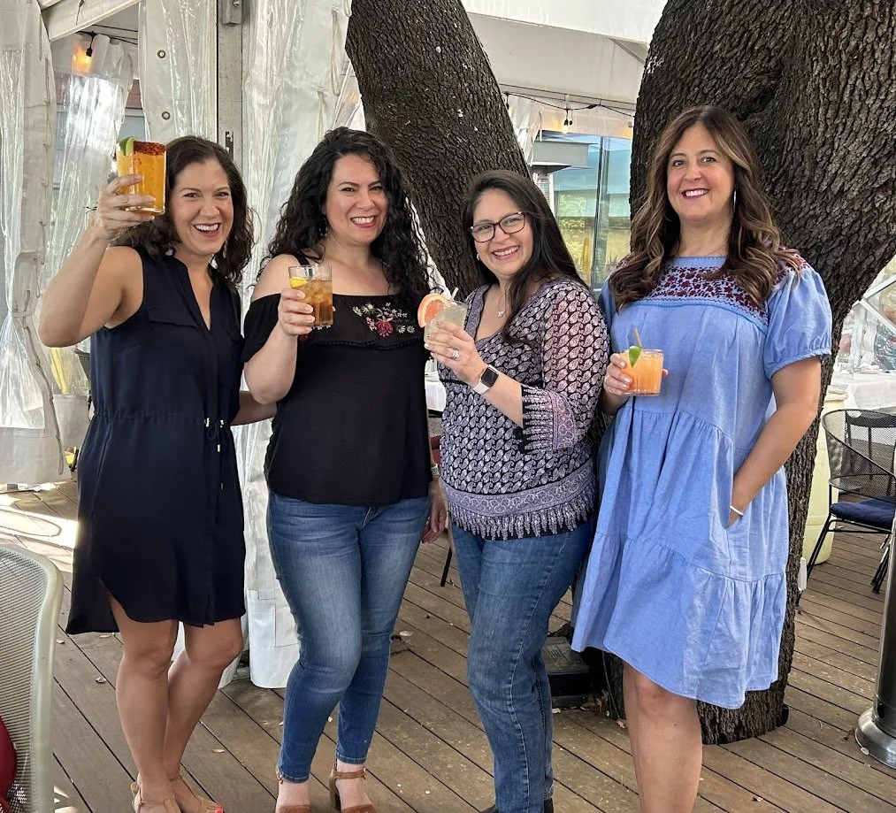 Four women smiling and holding drinks outdoors near large trees and a tented area.