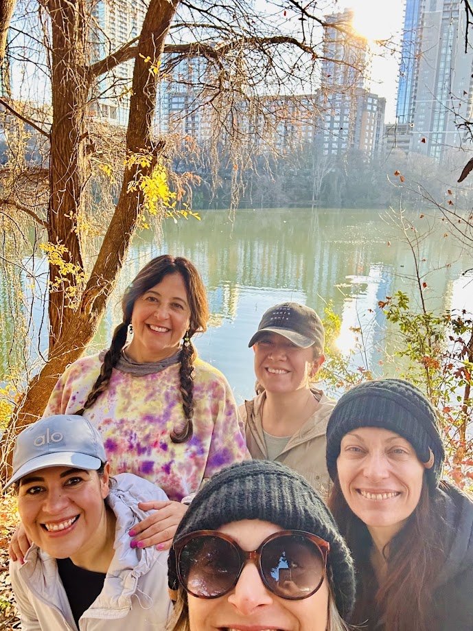 Group of five women outdoors by a lake with trees and city buildings in the background, smiling and taking a selfie.