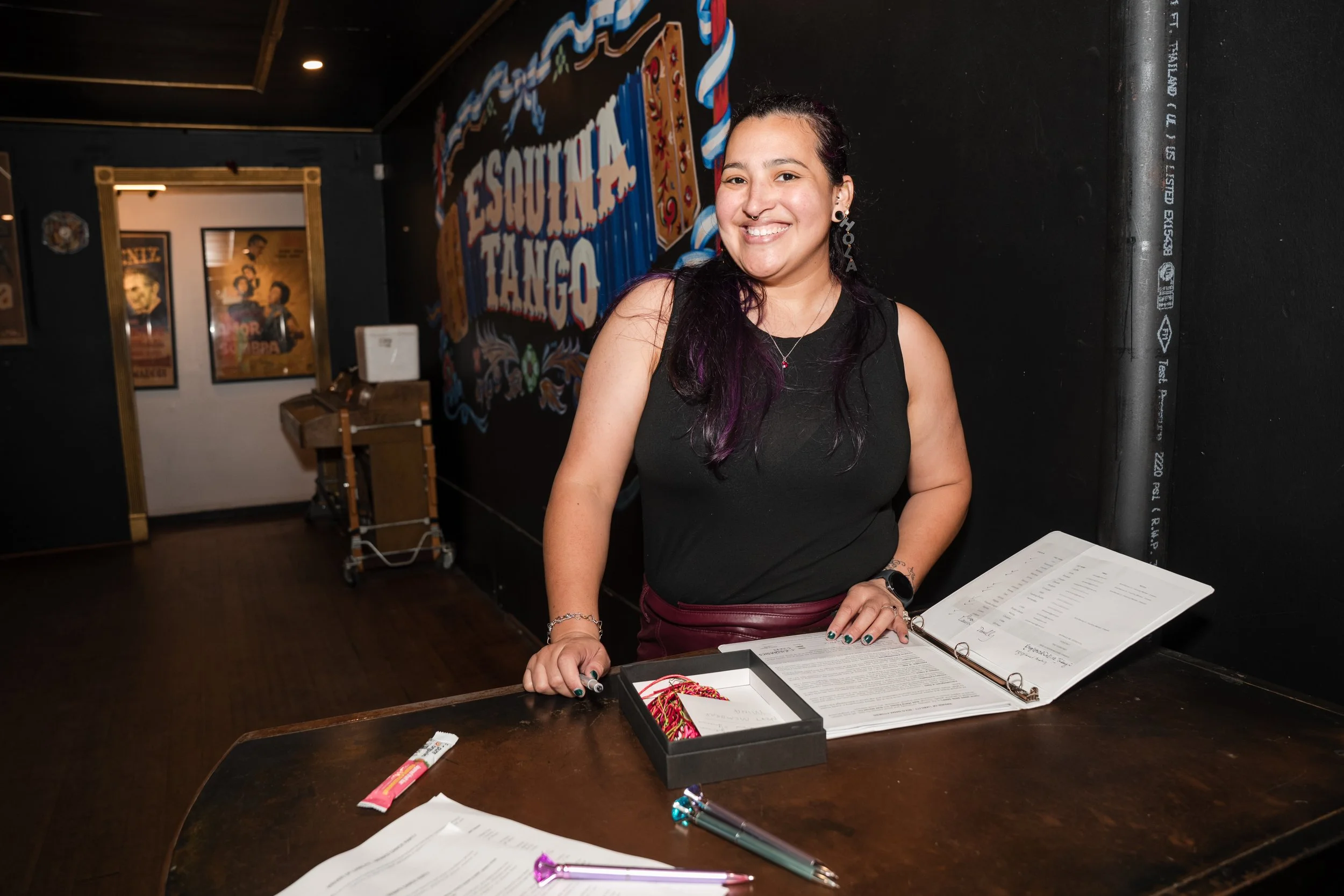 A woman with dark hair and a septum piercing smiling and standing behind a table with open sheet music. On the table are some pens, a box of colorful string, and a tube of candy. Behind her, a wall with a colorful sign and framed posters.