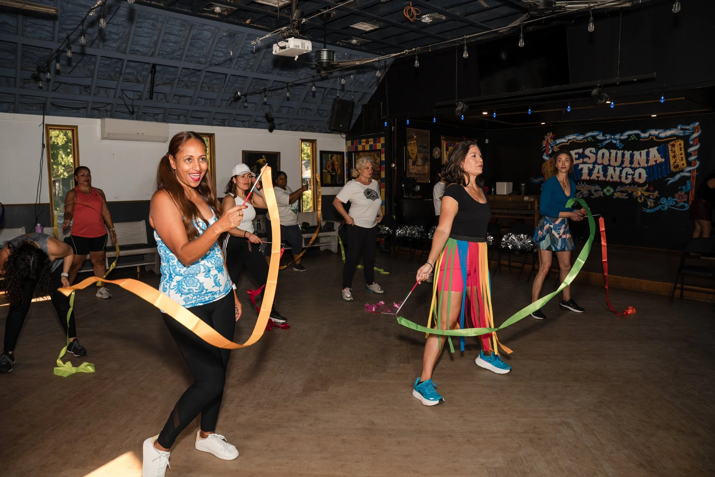 Women participating in a dance or exercise class indoors, using colorful ribbons, with a sign that reads "RESQUINA TANGO" in the background.