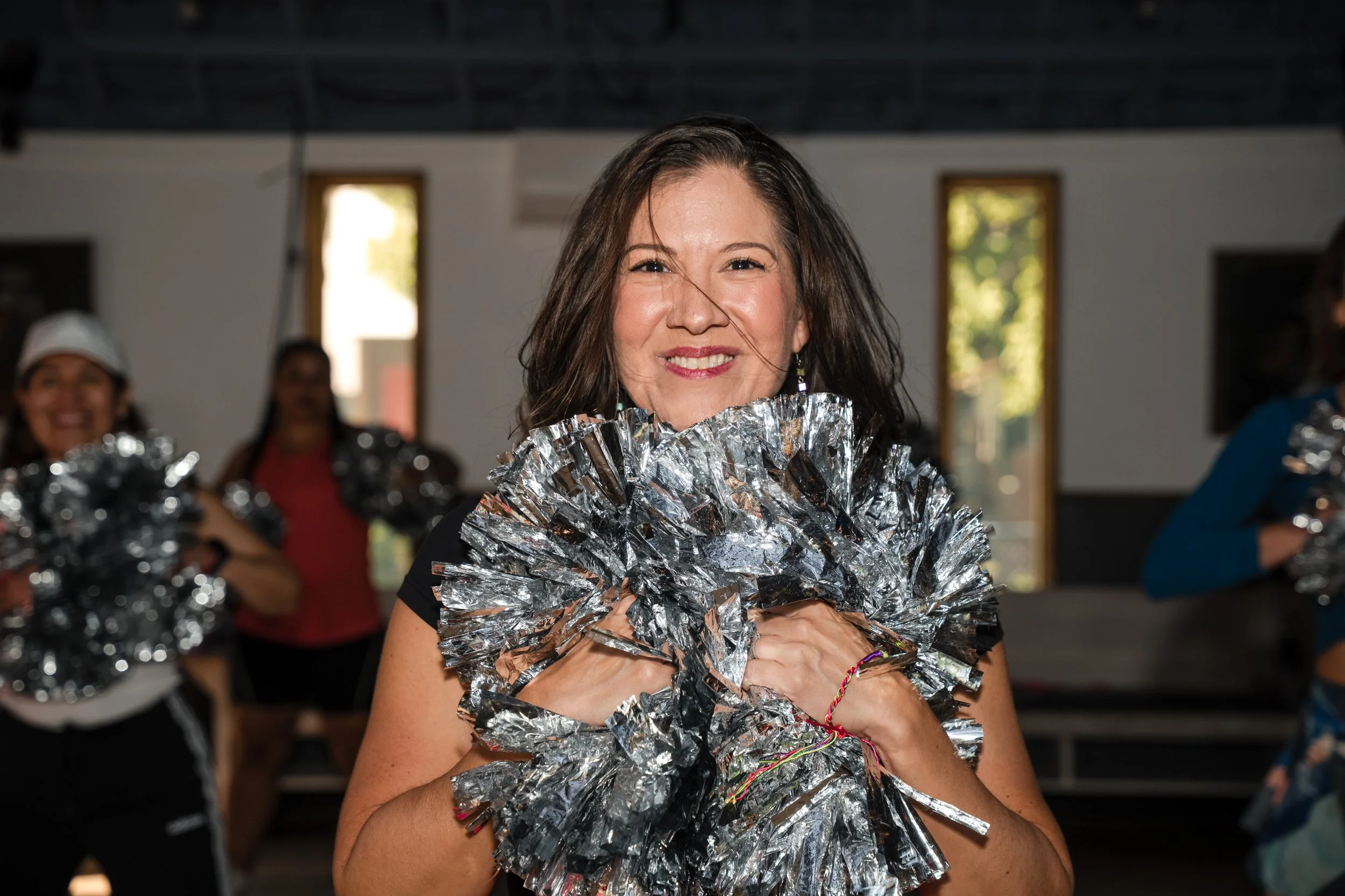 Melissa Acosta smiling holding metallic silver pom-poms, standing in room with open windows and other people in the background.