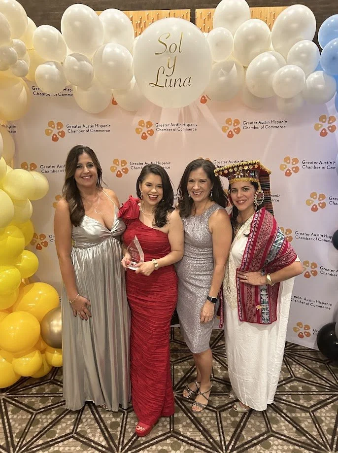 Four women dressed elegantly, standing together at an event with a backdrop that reads "Greater Austin" and balloons with a large centerpiece. One woman is wearing a traditional indigenous dress and headdress.