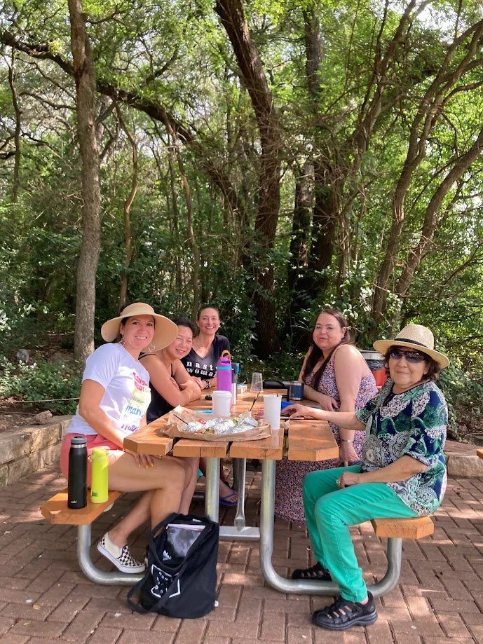 Five women sitting at a wooden picnic table outdoors surrounded by trees, enjoying a meal and drinks.