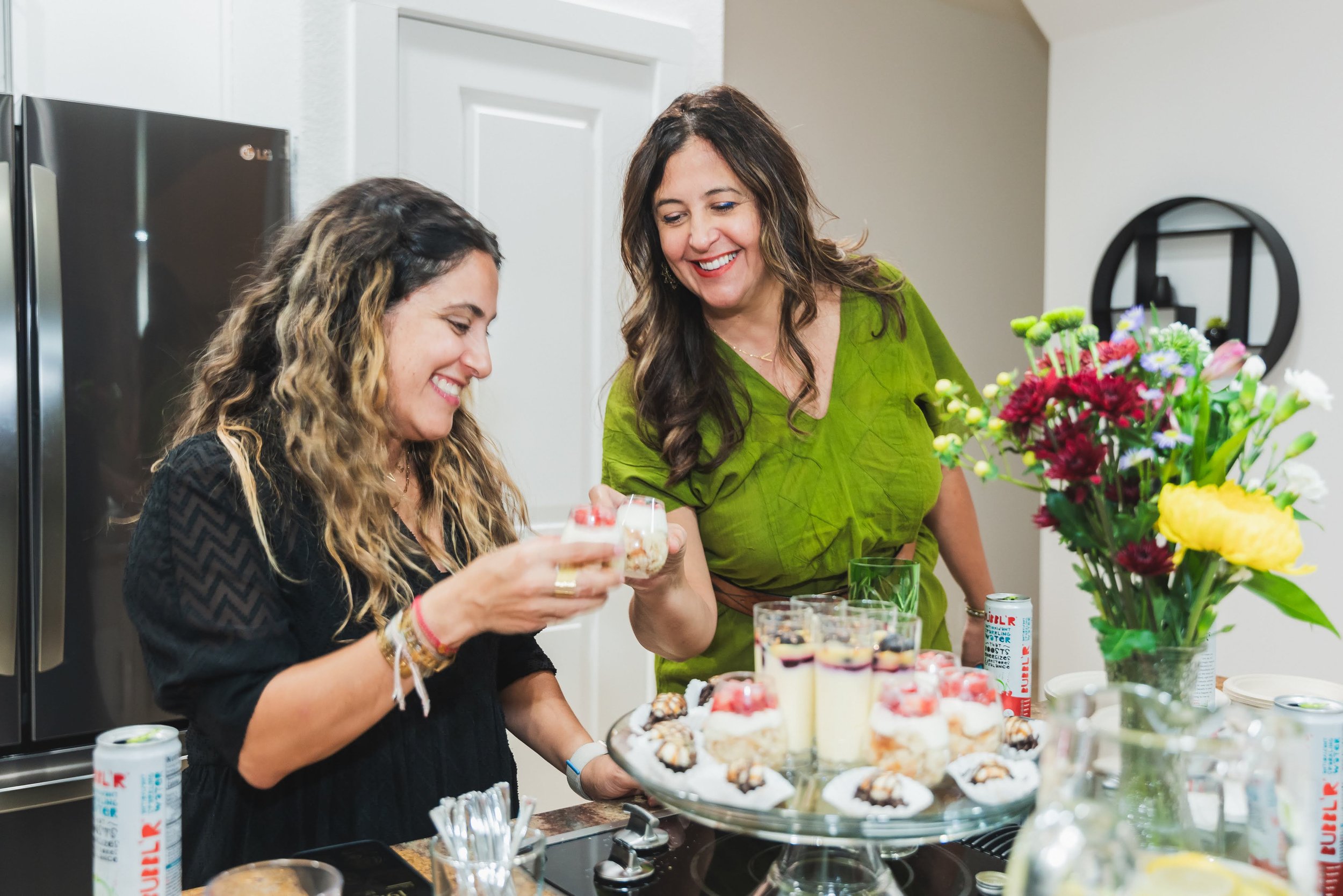 Two women smiling and chatting at a birthday bold mamas co. event, with a table of desserts and flowers in a kitchen.
