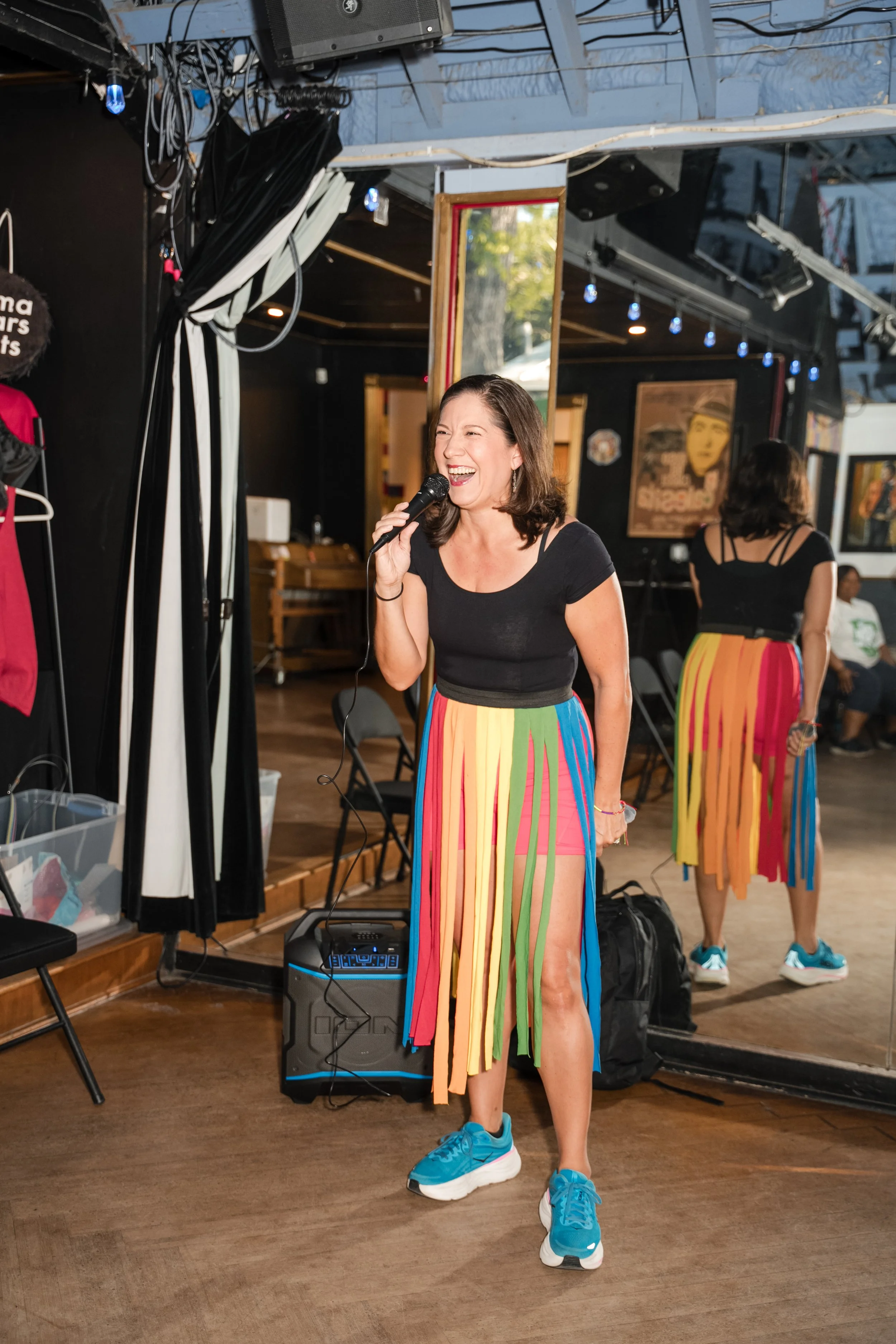 Woman smiling and speaking into a microphone while wearing a colorful rainbow fringe skirt, black top, and blue sneakers. She is standing in a room with a mirror, a black speaker on the floor, and various artworks on the walls.