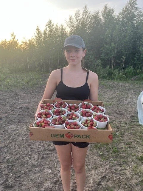 A young girl in a black tank top, shorts, and a baseball cap holding a tray of small bowls filled with strawberries outdoors during sunset, with trees in the background.