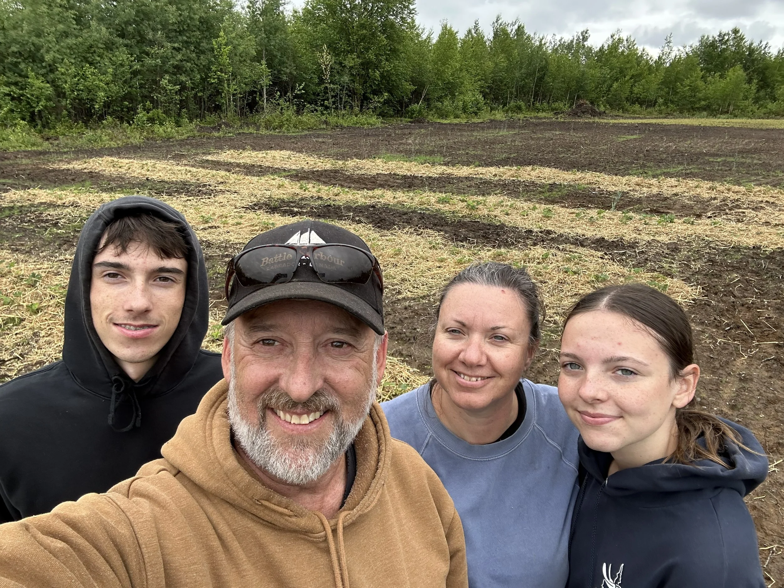 A family of four taking a selfie outdoors in front of a rural field with trees in the background.