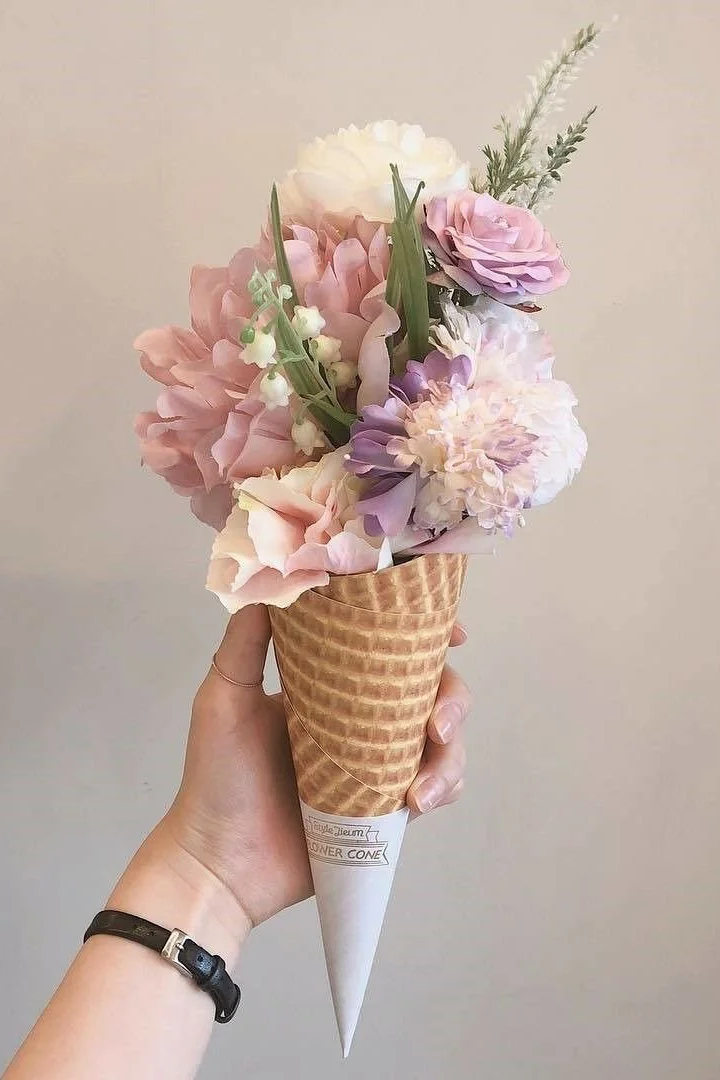 A person's hand holding a bouquet of pastel-colored flowers arranged in an ice cream cone.